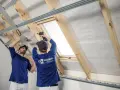 Dwóch mężczyzn montuje konstrukcję pod płyty gipsowo-kartonowe, przygotowując ocieplenie dachu. Jeden z nich używa poziomicy, drugi wkrętarki.
