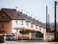 Rządowy dom szeregowy z jasnymi ścianami i ciemnym dachem, z białymi kominami. Przed domem ogrodzenie i samochód.