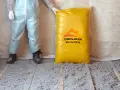 Osoba w kombinezonie stoi obok worka z materiałem do ocieplenia celulozą. Podłoga pokryta jest warstwą izolacji.