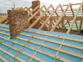 Drewniany dach jętkowy w budowie, z widoczną ceglaną kominową i niebieską membraną dachową.