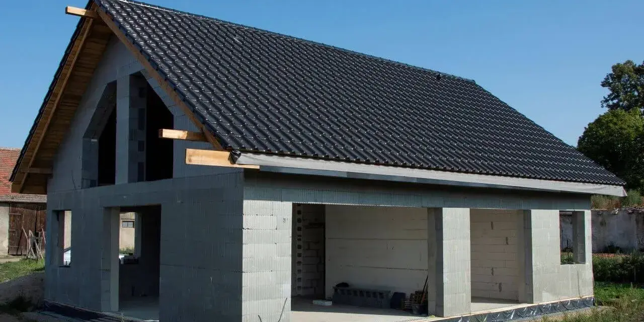 Dom w stanie surowym otwartym, z dachem pokrytym czarną dachówką. Ściany z bloczków betonowych, bez okien i drzwi.