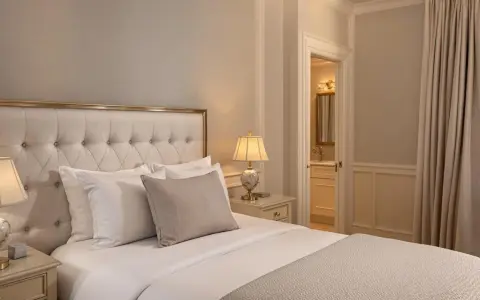 Elegancka sypialnia z pikowanym zagłówkiem, dwoma lampkami nocnymi i widokiem na łazienkę. Idealne miejsce dla wynajmującego szukającego komfortu.