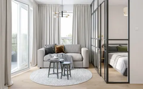 Jasna, nowoczesna kawalerka z wydzieloną sypialnią za szklaną ścianką. W salonie sofa, stoliki i okrągły dywan.