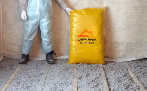 Osoba w kombinezonie stoi obok worka z materiałem do ocieplenia celulozą. Podłoga pokryta jest warstwą izolacji.