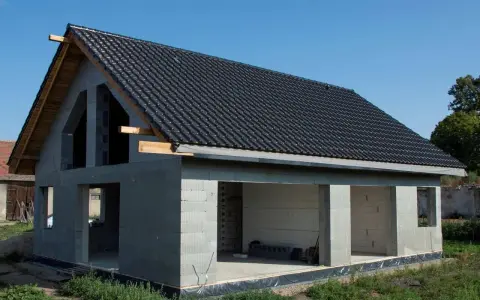 Dom w stanie surowym otwartym, z dachem pokrytym czarną dachówką. Ściany z bloczków betonowych, bez okien i drzwi.