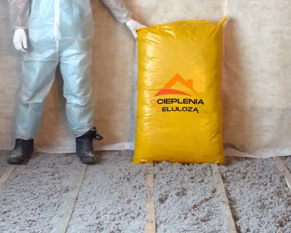 Osoba w kombinezonie stoi obok worka z materiałem do ocieplenia celulozą. Podłoga pokryta jest warstwą izolacji.