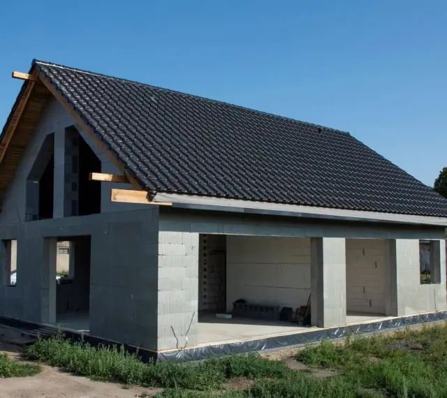 Dom w stanie surowym otwartym, z dachem pokrytym czarną dachówką. Ściany z bloczków betonowych, bez okien i drzwi.