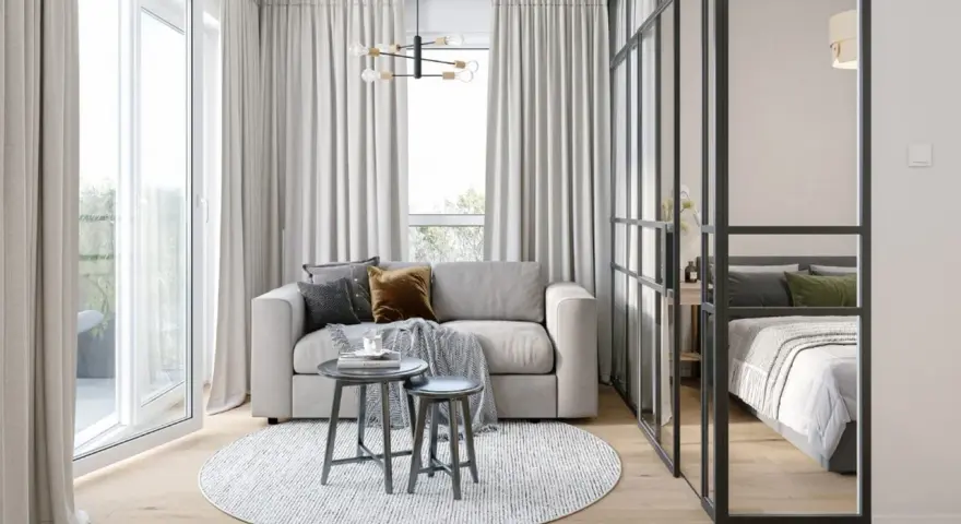 Jasna, nowoczesna kawalerka z wydzieloną sypialnią za szklaną ścianką. W salonie sofa, stoliki i okrągły dywan.