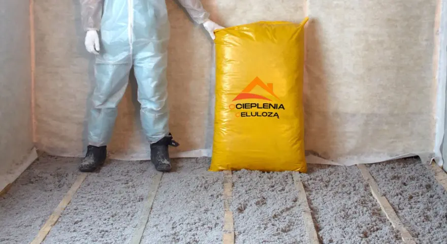 Osoba w kombinezonie stoi obok worka z materiałem do ocieplenia celulozą. Podłoga pokryta jest warstwą izolacji.