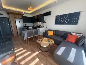 Nowoczesny salon z aneksem kuchennym. Wygodna kanapa, stolik kawowy, stół z krzesłami. W tle lodówka i sprzęty kuchenne. Idealne miejsce na relaks, z myślą o komforcie gości.
