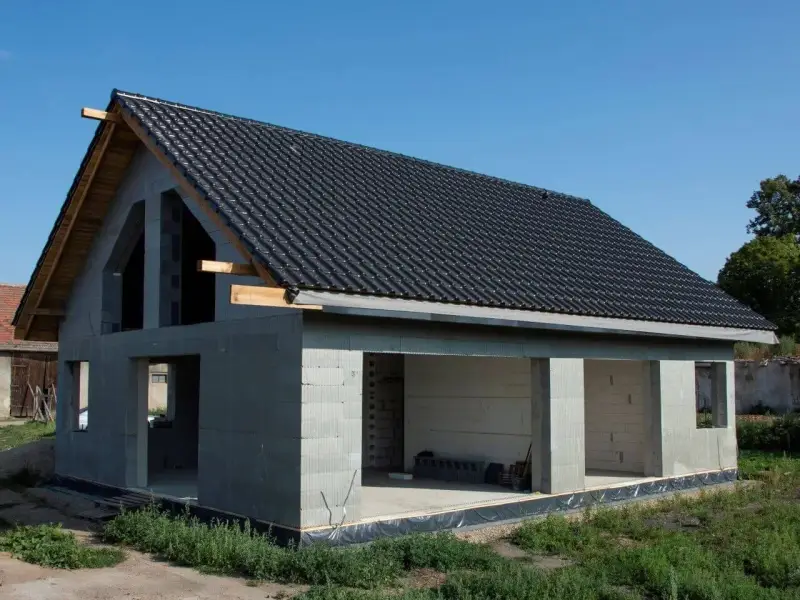 Dom w stanie surowym otwartym, z dachem pokrytym czarną dachówką. Ściany z bloczków betonowych, bez okien i drzwi.